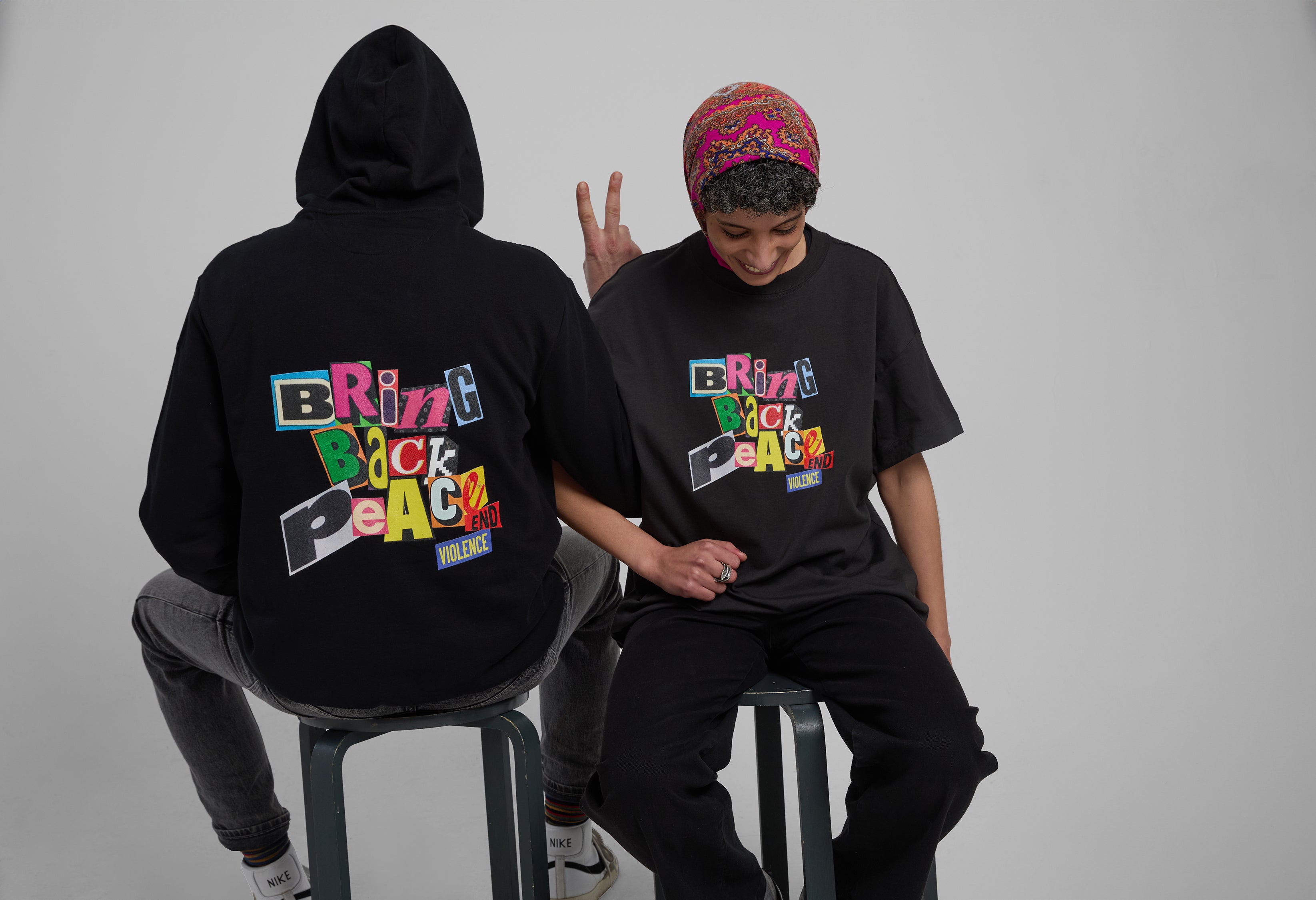 Two people sitting on stools wearing hoodies with colorful text.