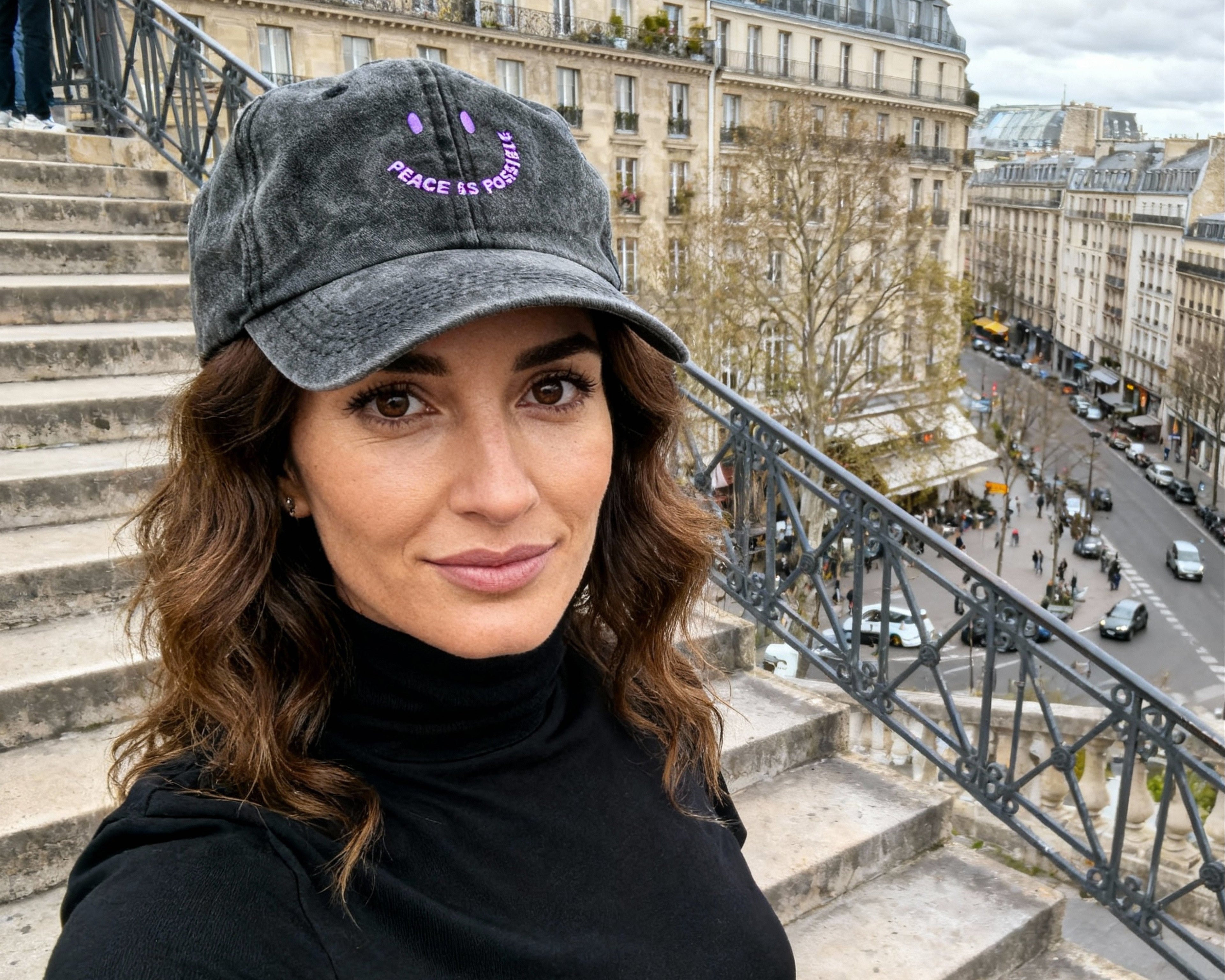 Woman wearing a black turtleneck and gray cap on stone steps with a cityscape background